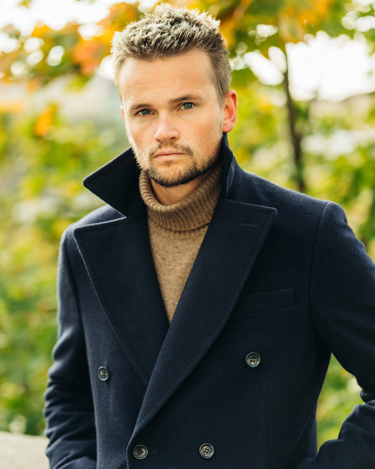Man wearing a dark coat and brown turtleneck sweater outdoors with blurred greenery in the background