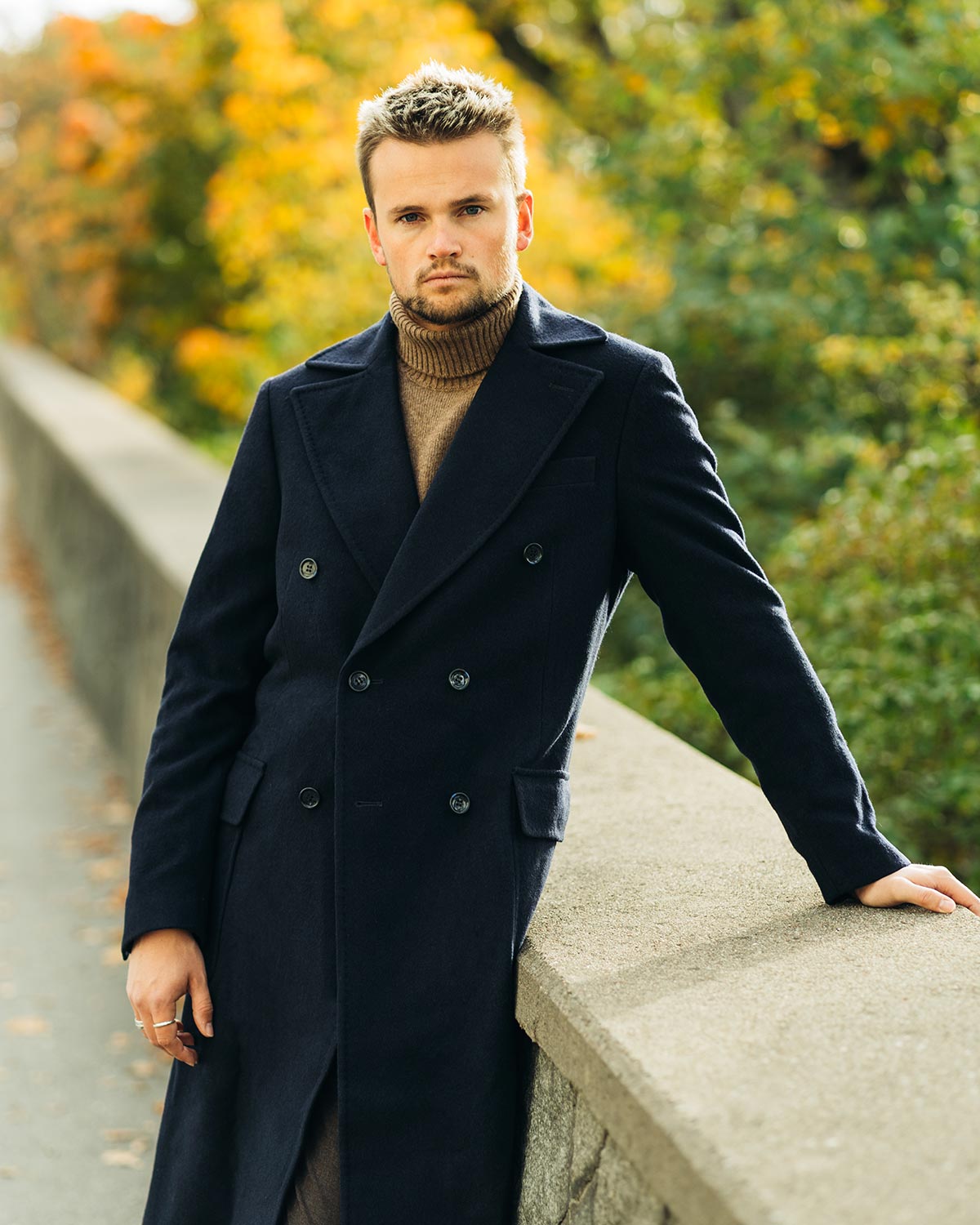 Man in a dark coat standing on a stone wall with autumn trees in the background