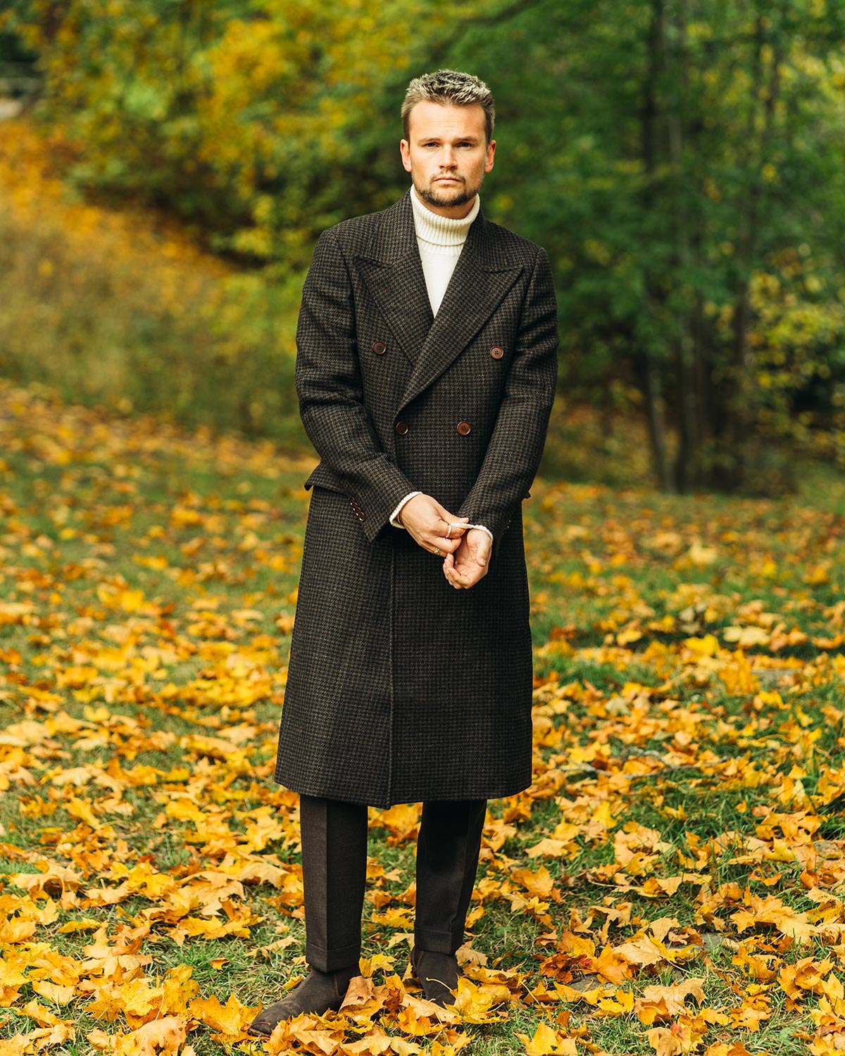 Man wearing a brown double breasted overcoat standing in an autumnal park with fallen leaves and trees in the background.