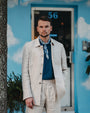 Man wearing a beige jacket and blue shirt standing in front of a blue wall with white cloud patterns.