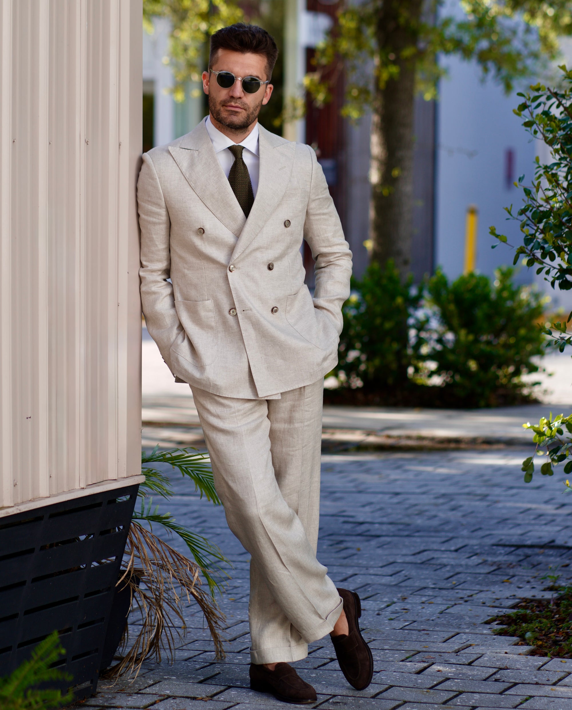 Man in a beige suit walking outdoors with plants and a building in the background