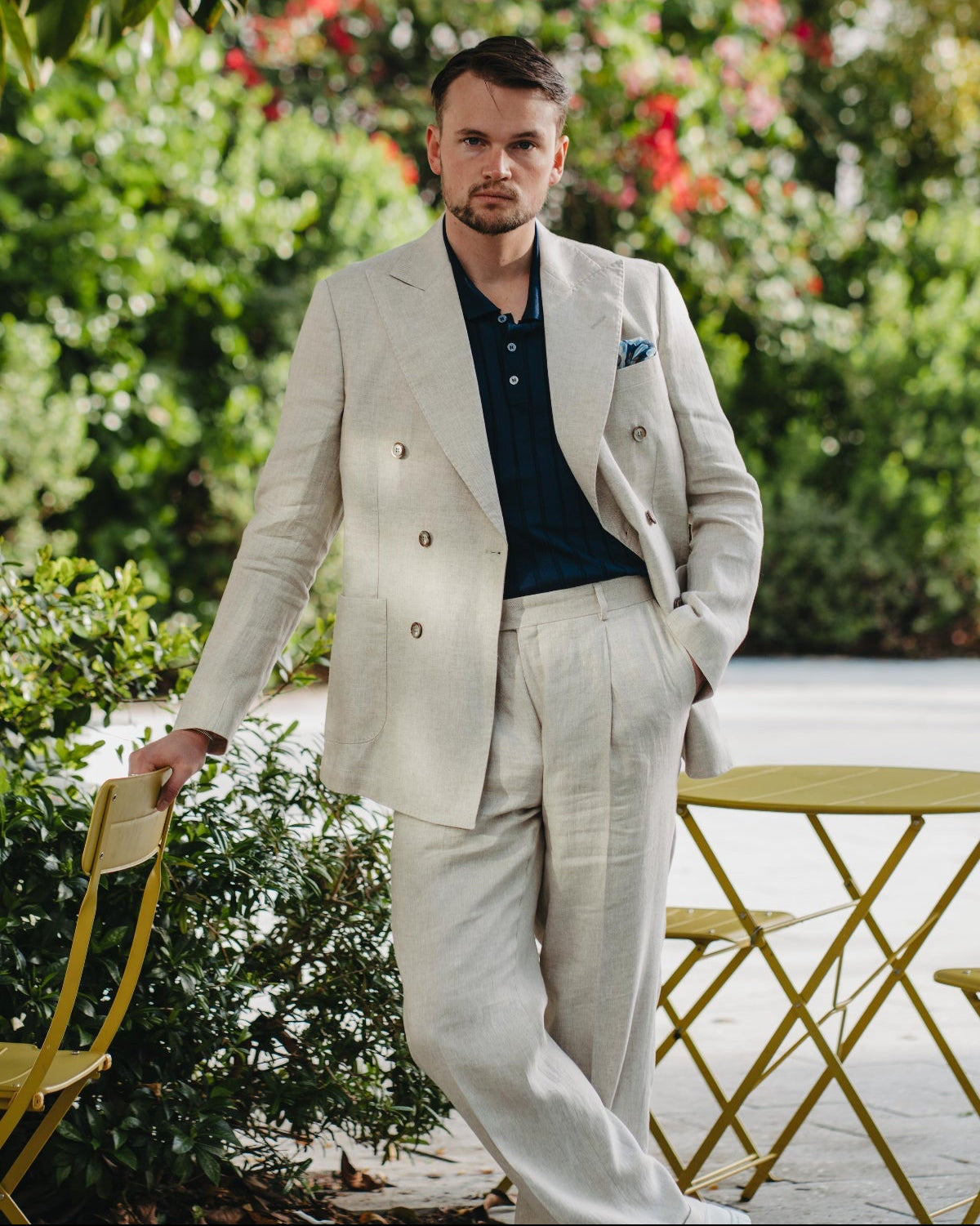 Man in a light-colored suit standing outdoors with greenery and flowers in the background