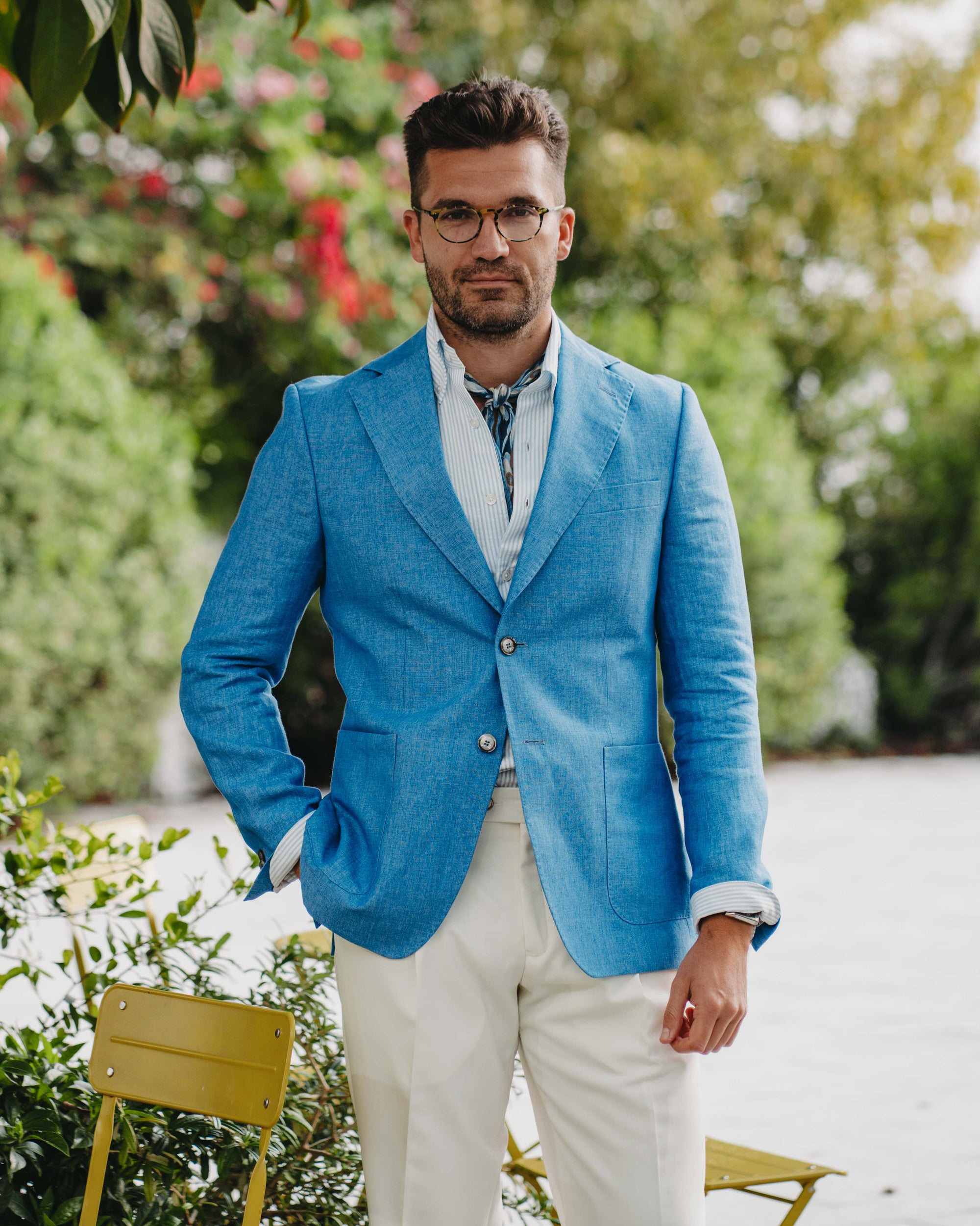 Man wearing a blue blazer and white pants standing outdoors with greenery in the background