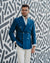 Man wearing a blue blazer and white pants standing against a patterned wall.