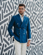 Man wearing a blue blazer and white pants standing against a patterned wall.
