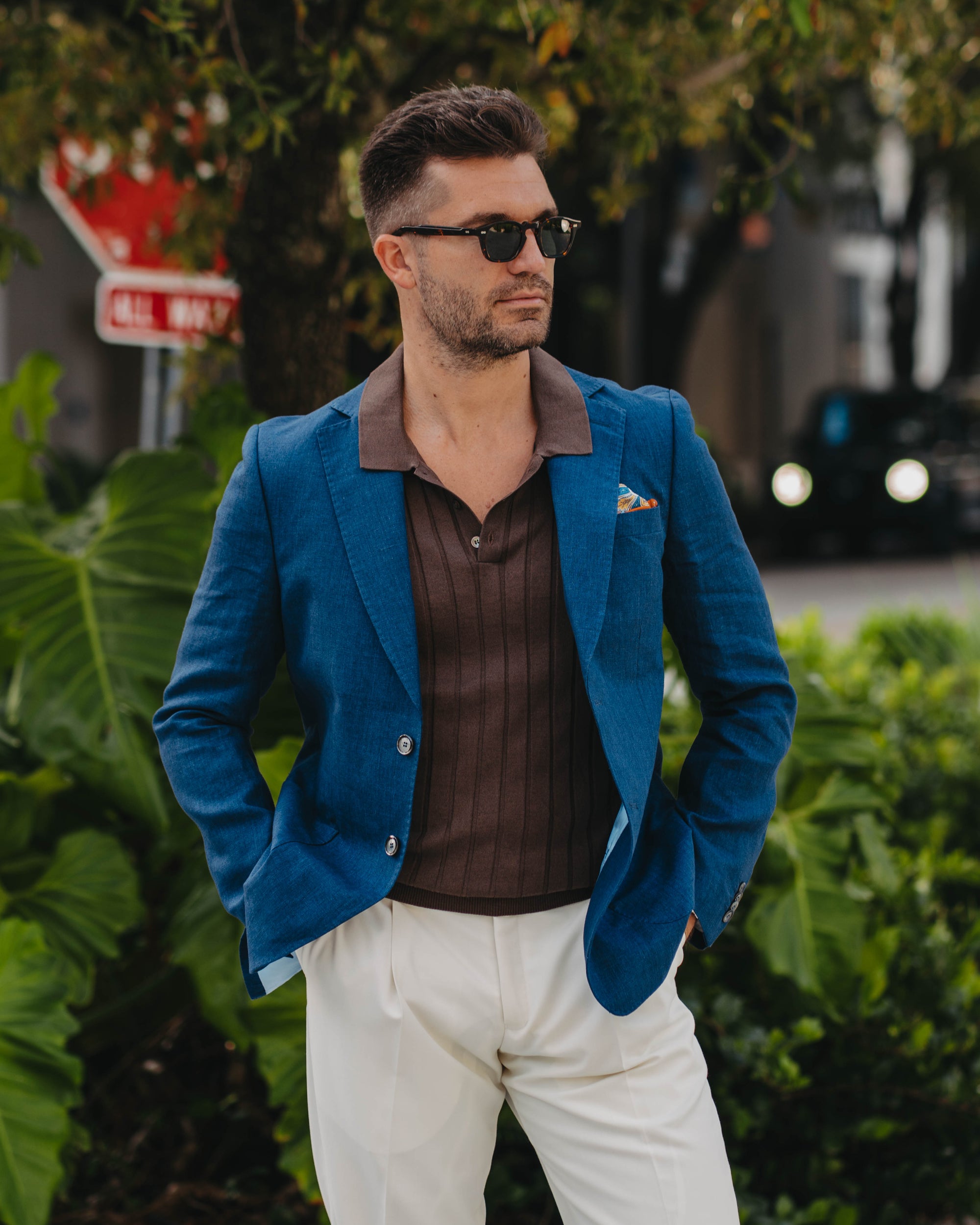 Man wearing a blue blazer over a brown shirt with white pants, standing outdoors.