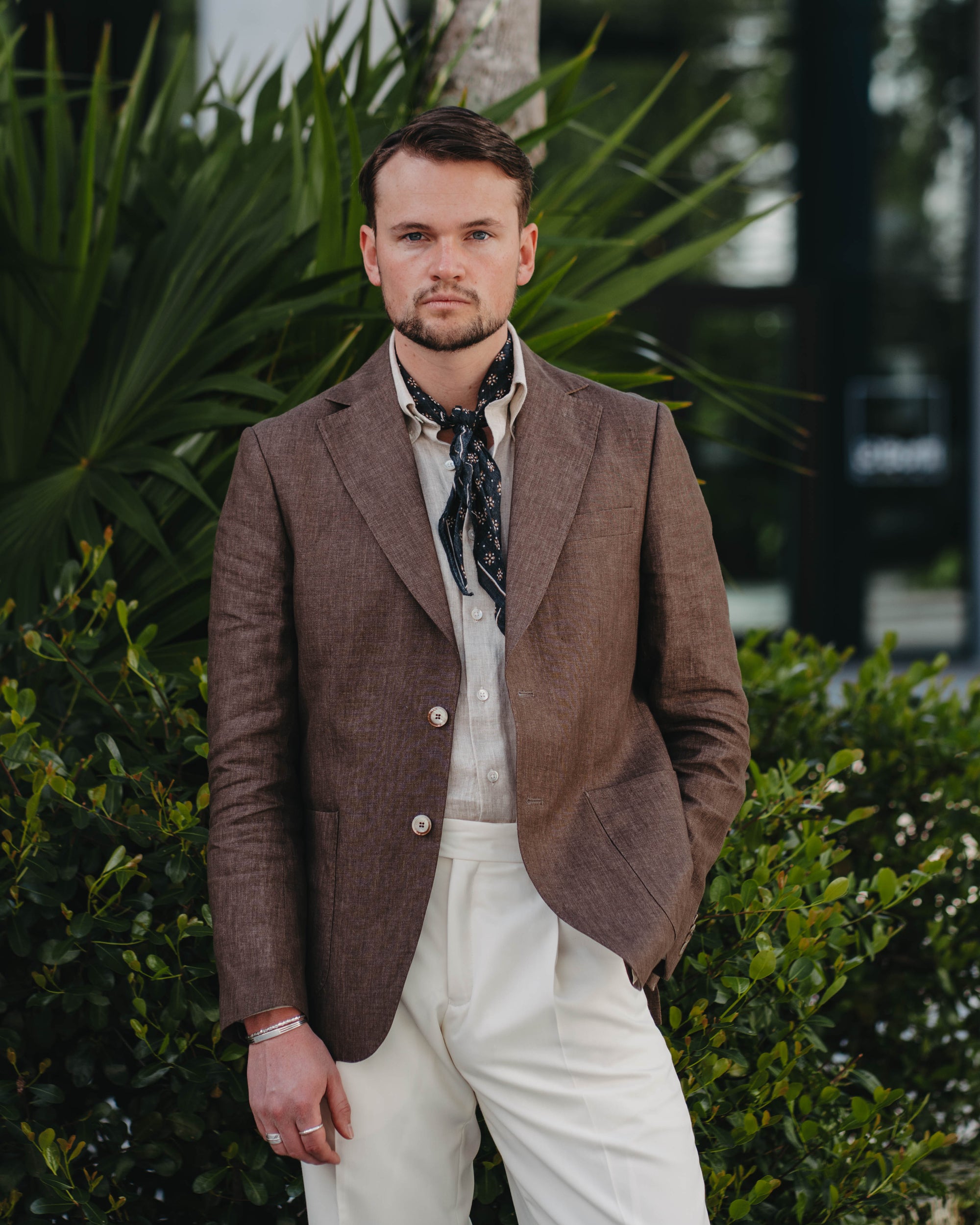 Man wearing a brown blazer and white pants standing outdoors with greenery in the background