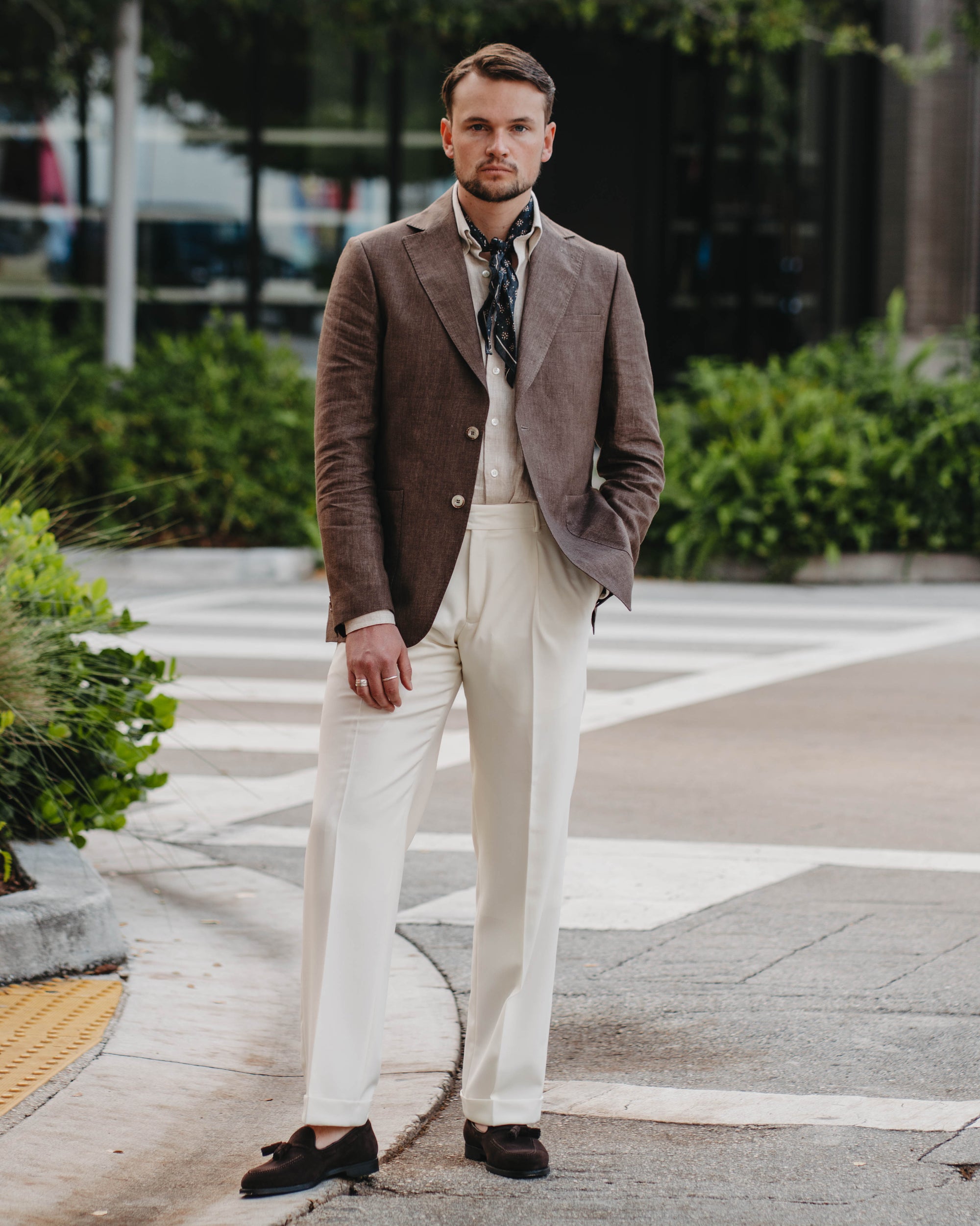 Man in a brown blazer and white pants standing on a sidewalk with greenery in the background