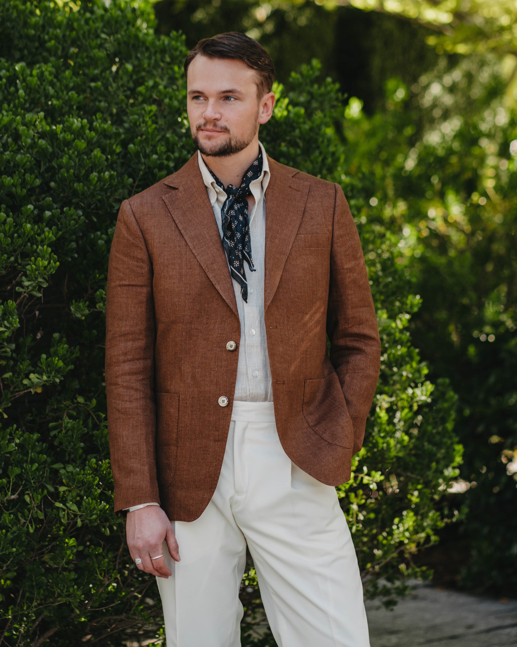 Man wearing a brown blazer and white pants standing in front of green foliage.