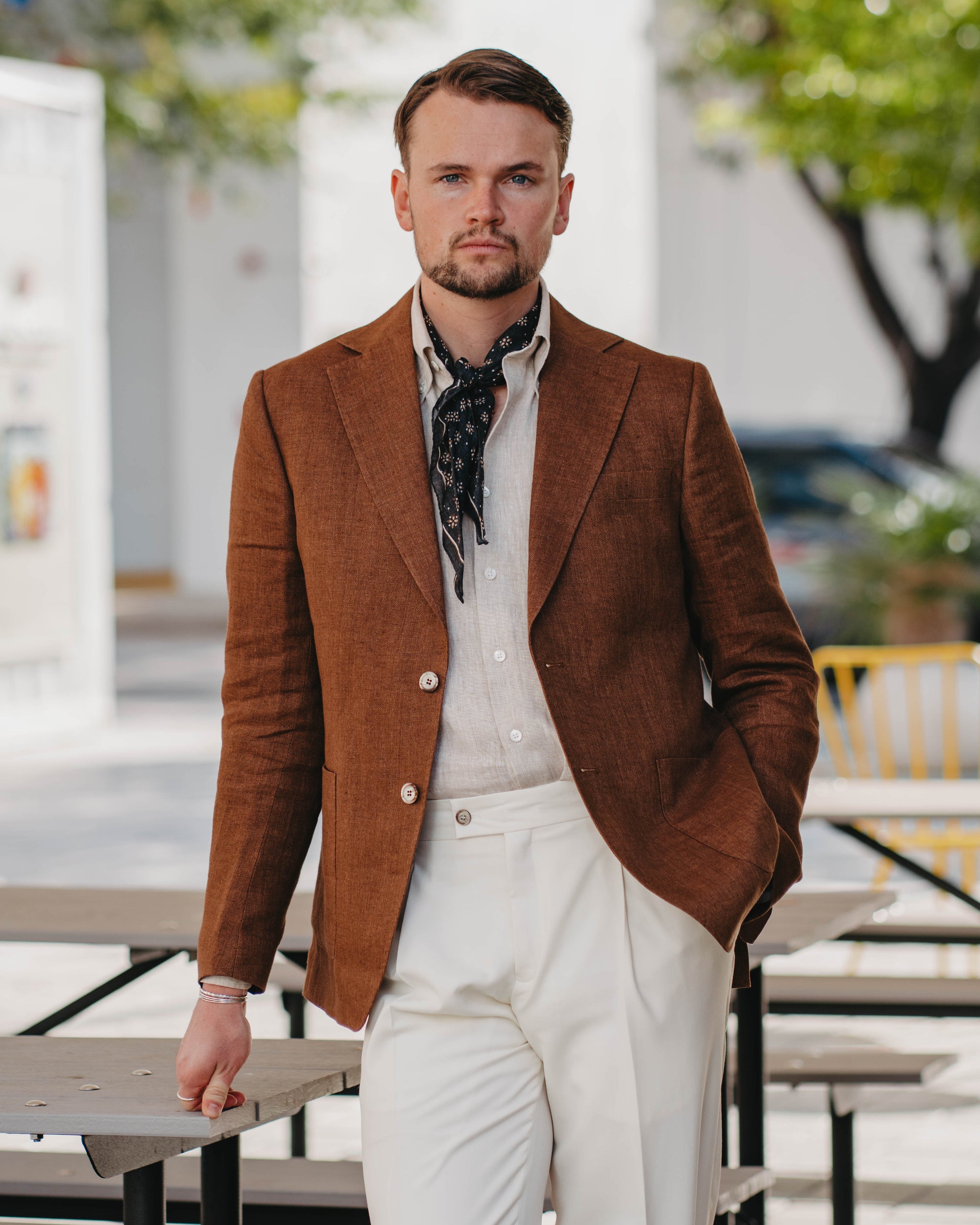 Man wearing a brown blazer over a beige shirt with a patterned scarf, standing outdoors.