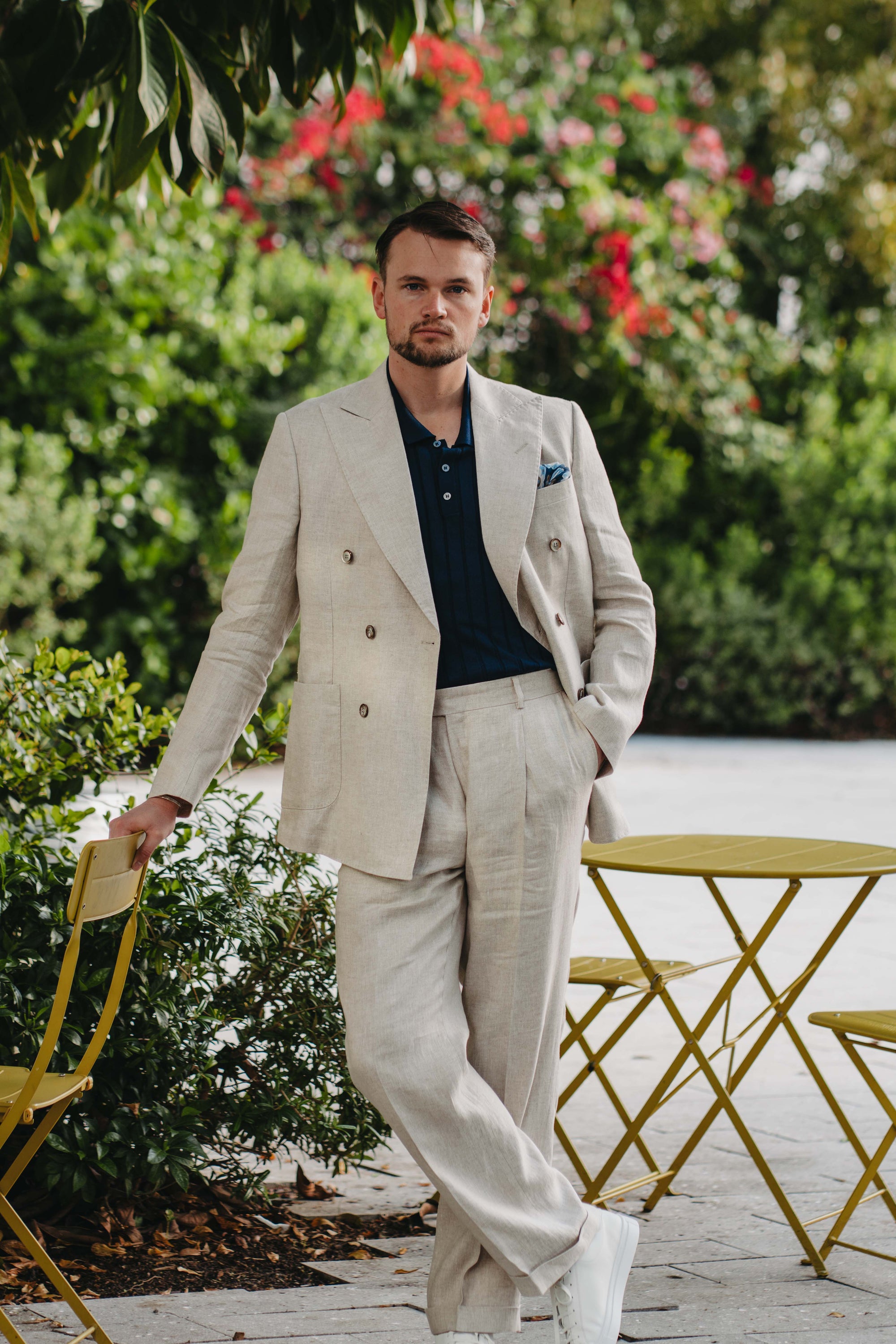 Man in a beige suit standing and blue sweater outdoors with greenery and flowers in the background