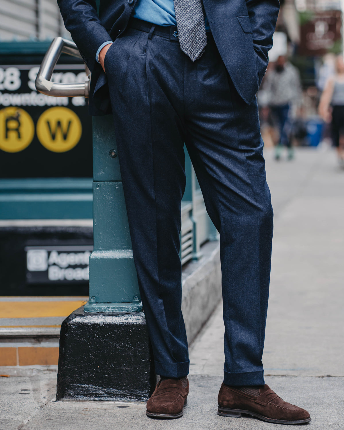 A man wearing a navy blue suit, light blue dress shirt, patterned tie, and brown suede loafers, standing on a city sidewalk next to a subway station entrance.