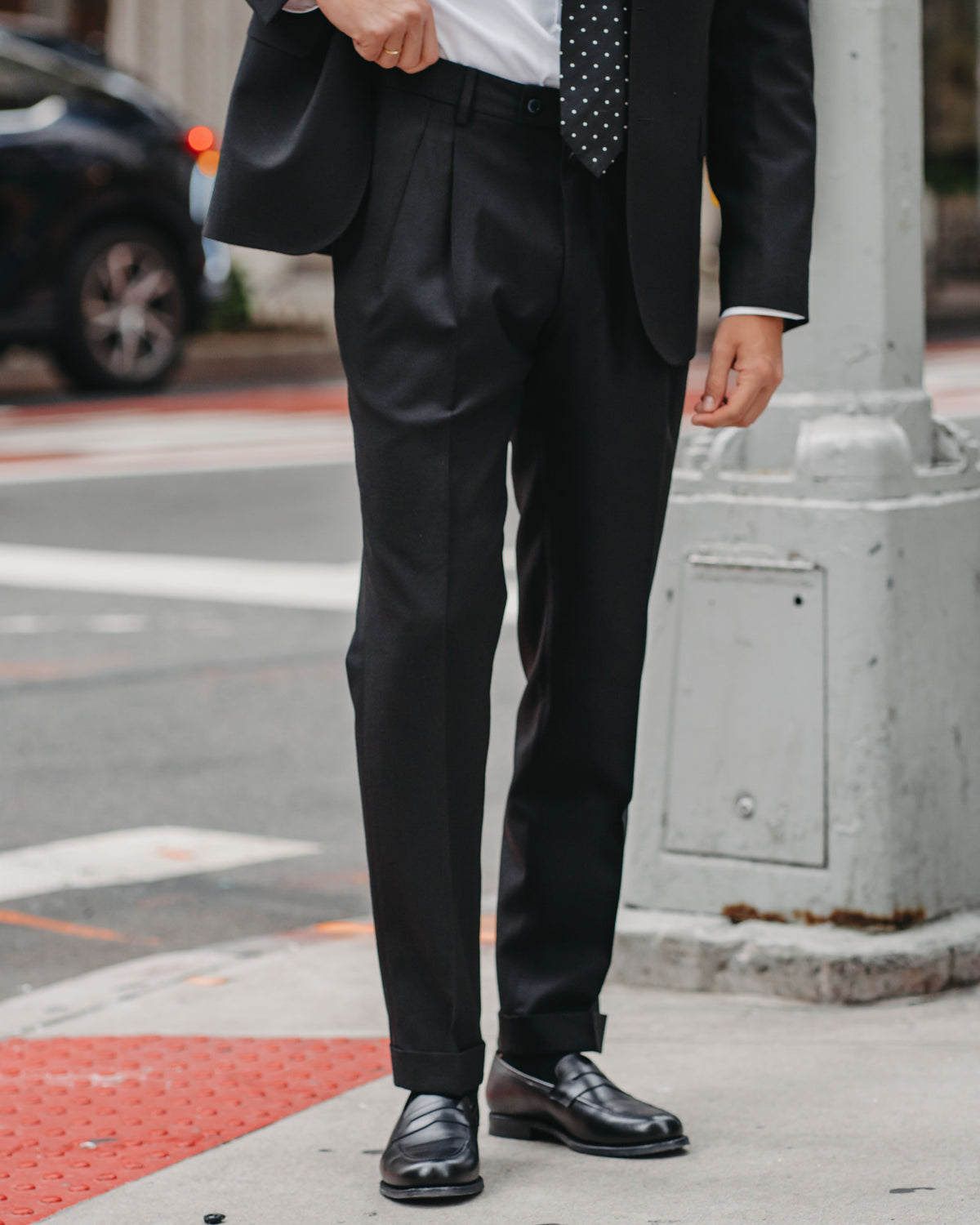 A man wearing a black suit, white dress shirt, and a black polka-dot tie, standing on a city sidewalk next to a street pole.