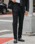 A man wearing a black suit, white dress shirt, and a black polka-dot tie, standing on a city sidewalk next to a street pole.