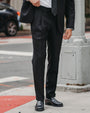 A man wearing a black suit, white dress shirt, and a black polka-dot tie, standing on a city sidewalk next to a street pole.