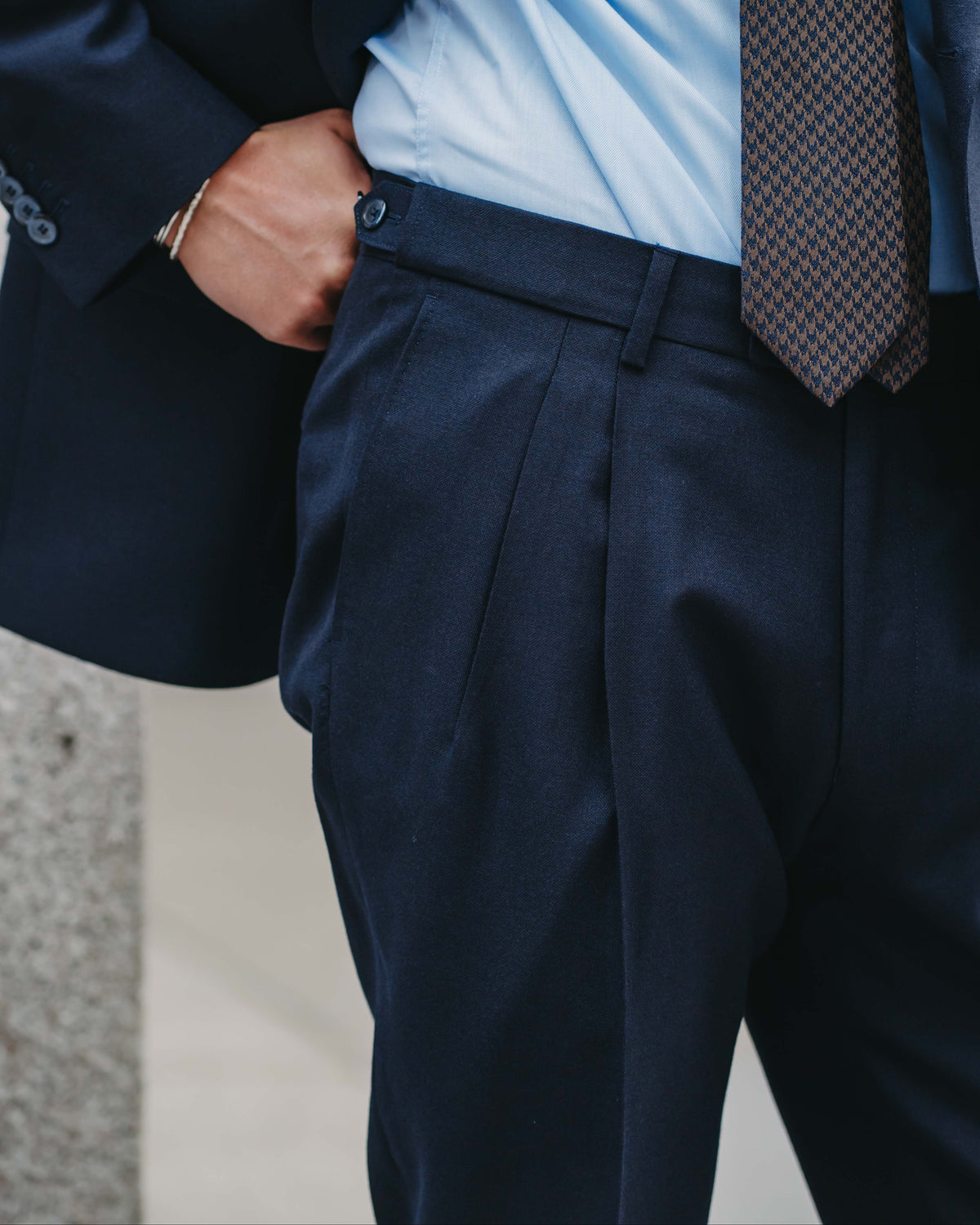 Close-up of a man wearing navy blue dress pants, a light blue dress shirt, and a brown patterned tie, with his hand in his pocket.