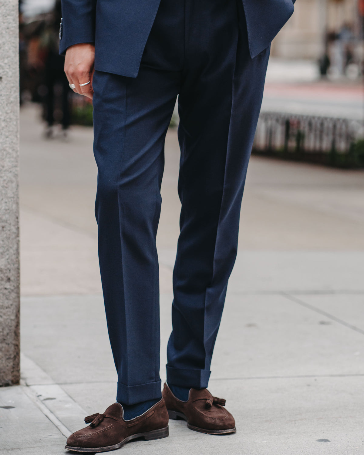 A man wearing a navy blue suit and brown suede tassel loafers, standing on a city sidewalk.