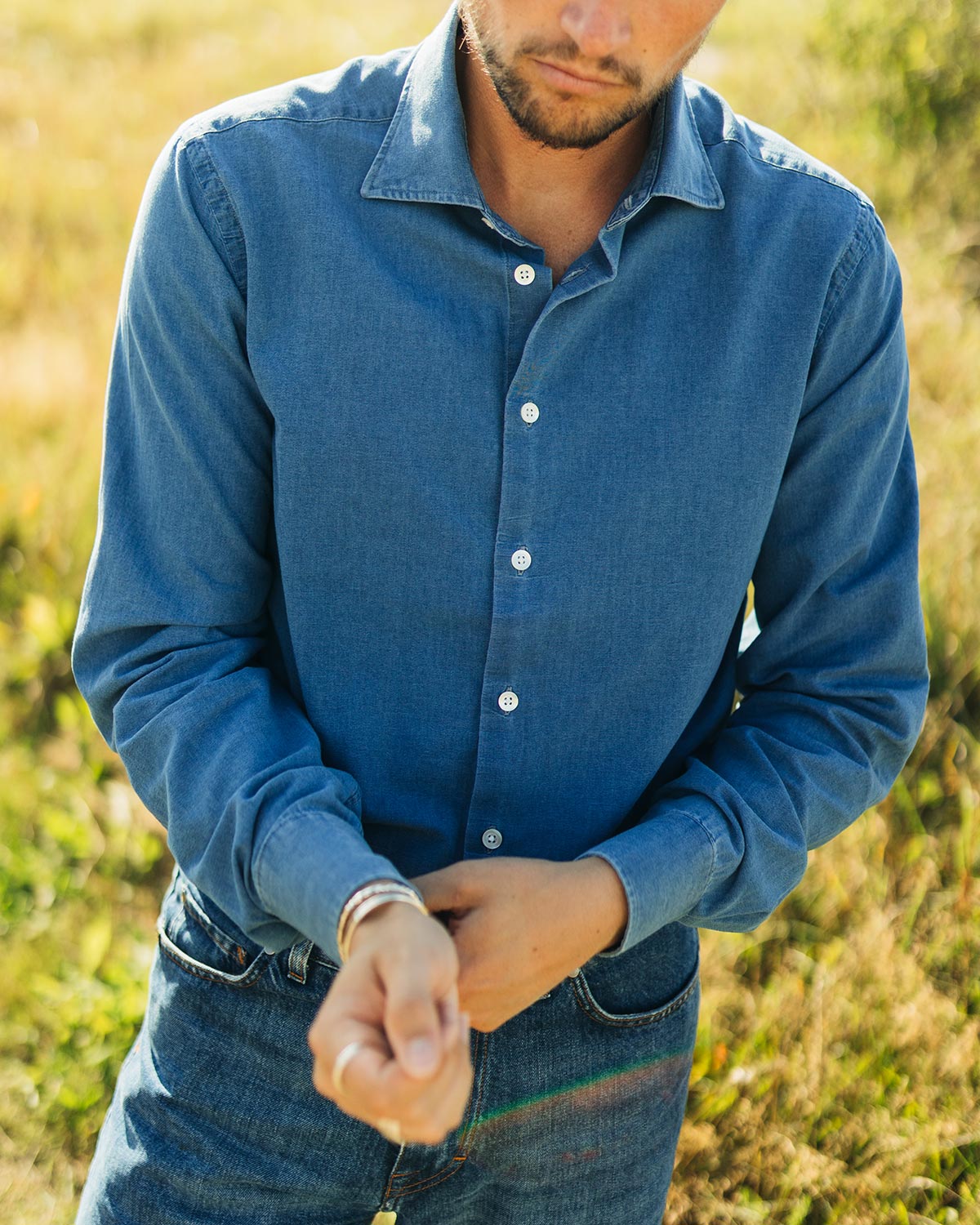 Man wearing a blue denim shirt and jeans standing in a grassy field.