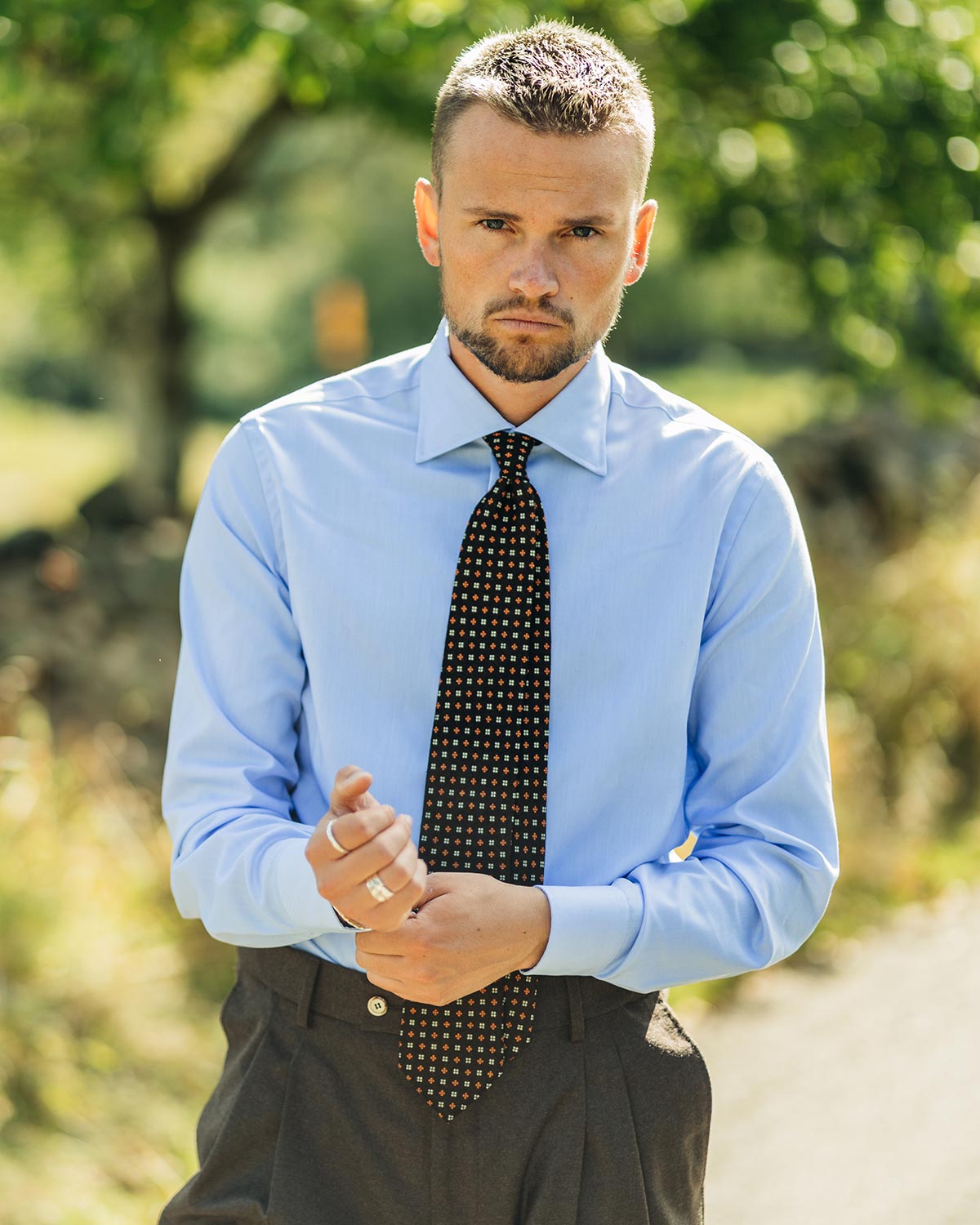 Man wearing a light blue shirt and dark tie outdoors with greenery in the background