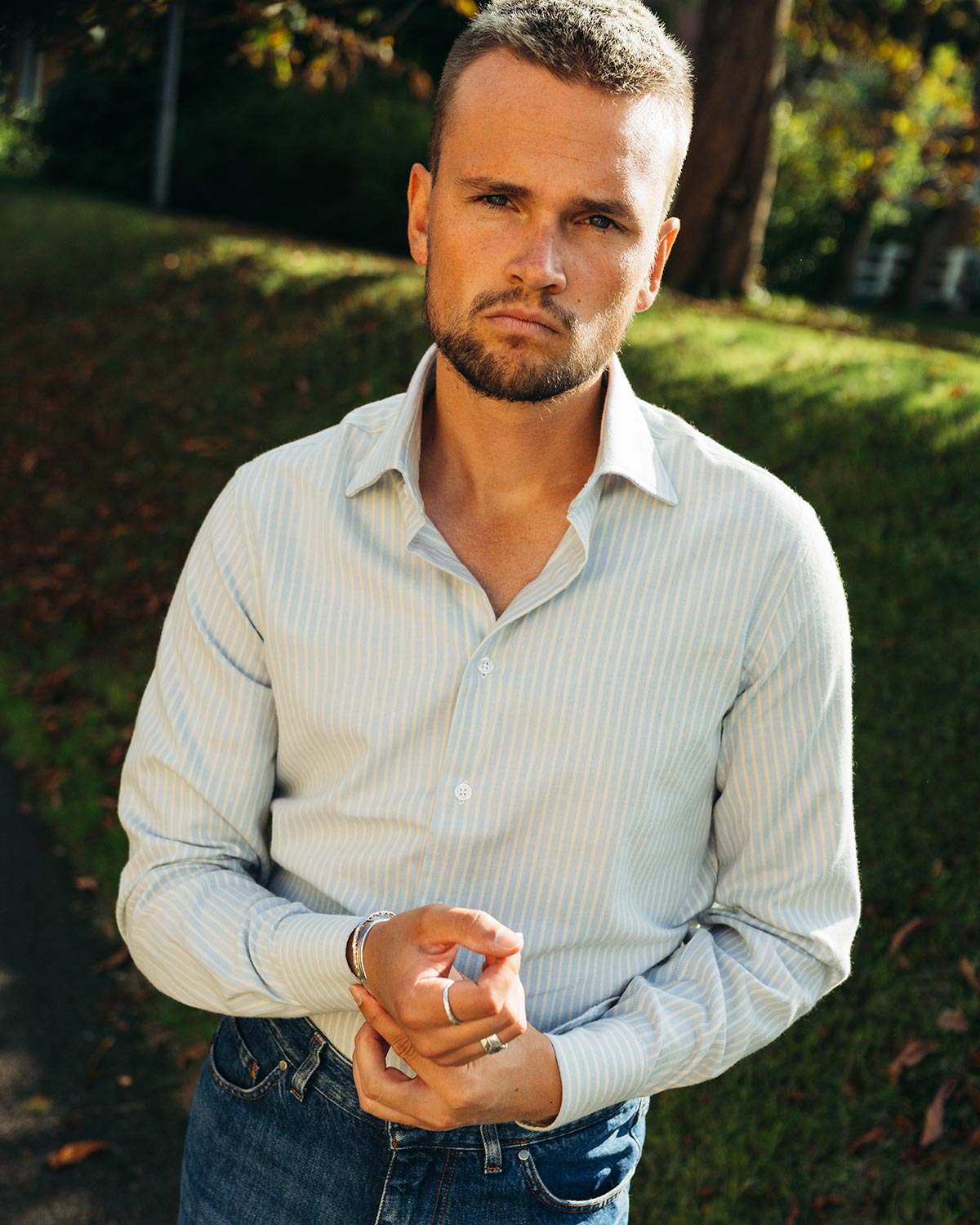 Man wearing a light blue striped flannel shirt and jeans standing outdoors with greenery in the background