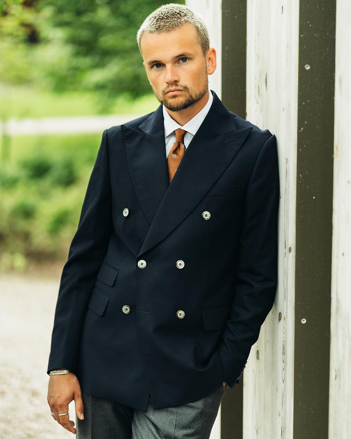 Man wearing a dark blue double-breasted suit with a patterned tie, standing against a blurred outdoor background.
