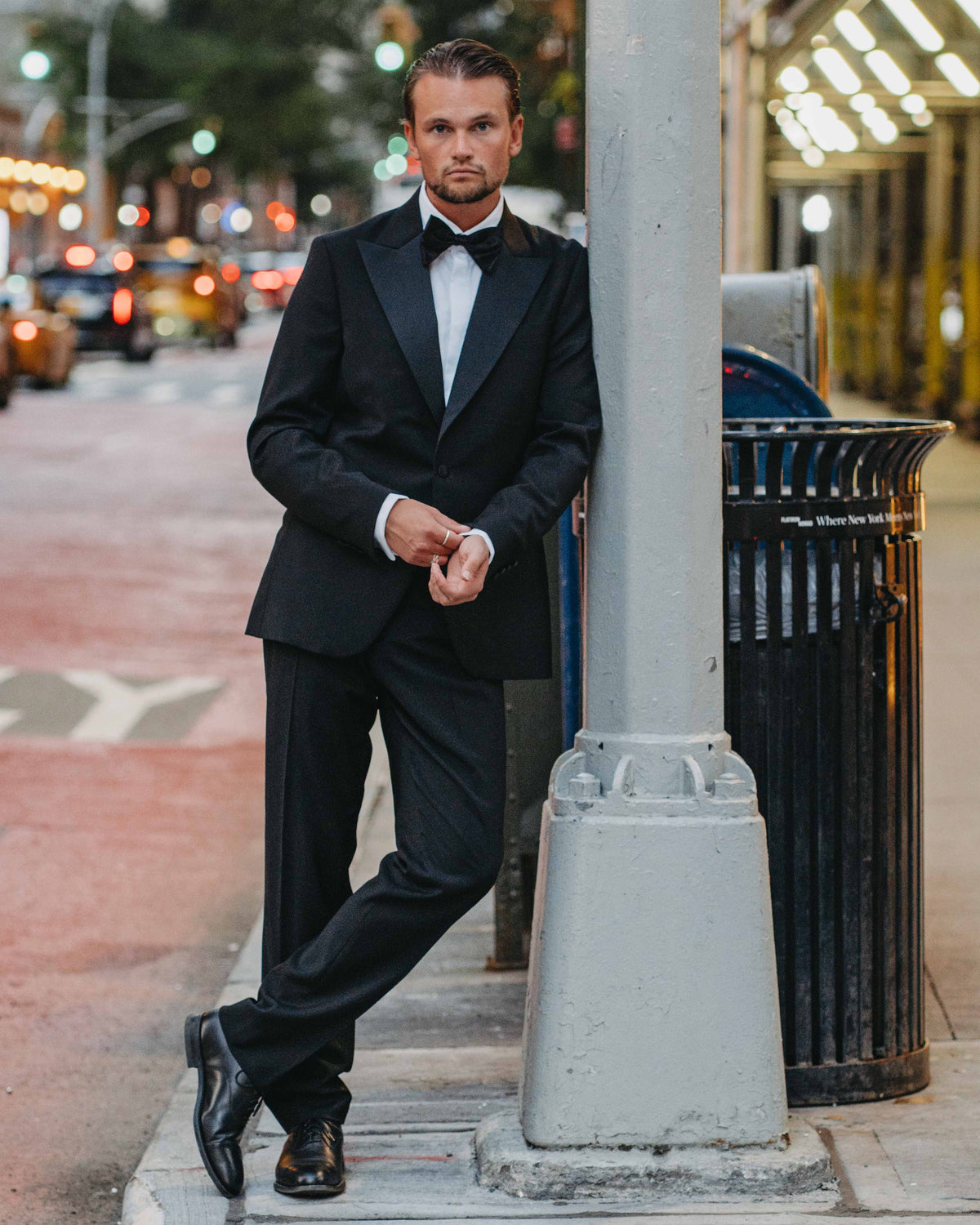 A man wearing a classic black tuxedo with a white shirt and black bow tie, leaning against a gray city street pole on a sidewalk.