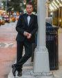 A man wearing a classic black tuxedo with a white shirt and black bow tie, leaning against a gray city street pole on a sidewalk.