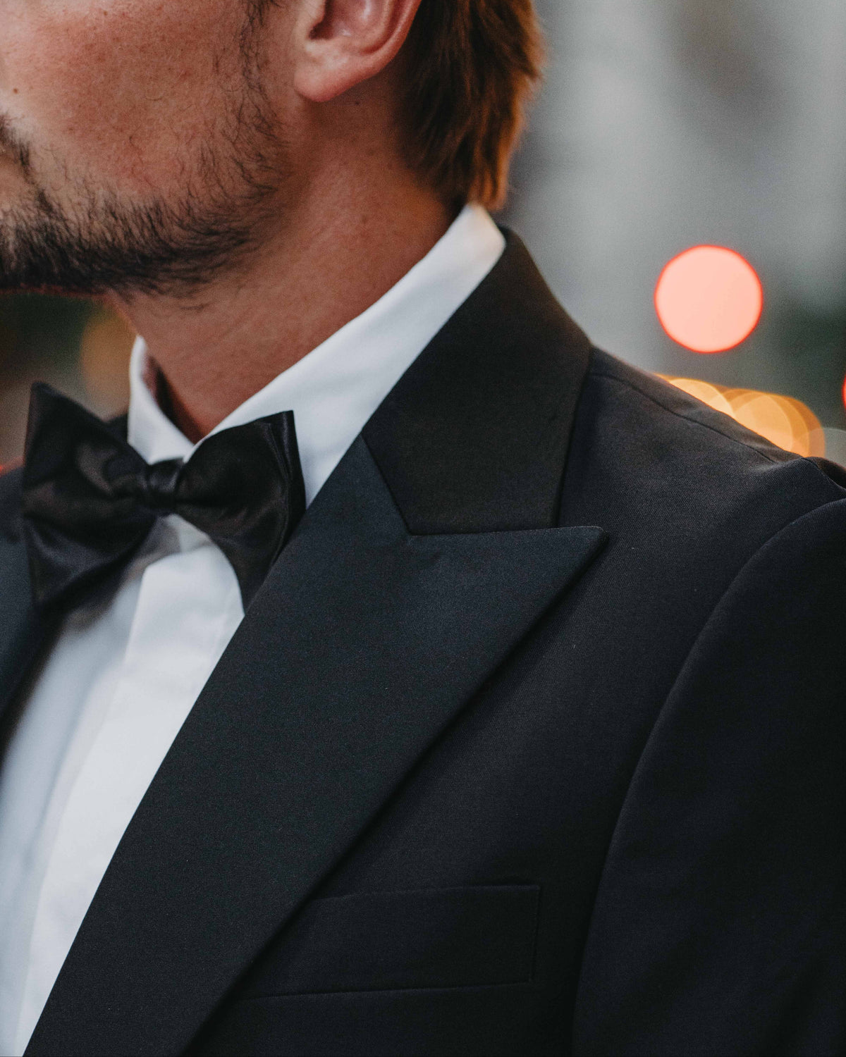A close-up side profile of a man wearing a classic black tuxedo jacket with satin peak lapels, a white dress shirt, and a black bow tie.