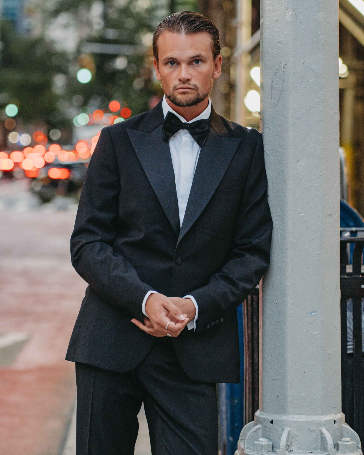 A man wearing a black tuxedo with a white dress shirt and black bow tie stands on a city street, leaning against a gray pole.