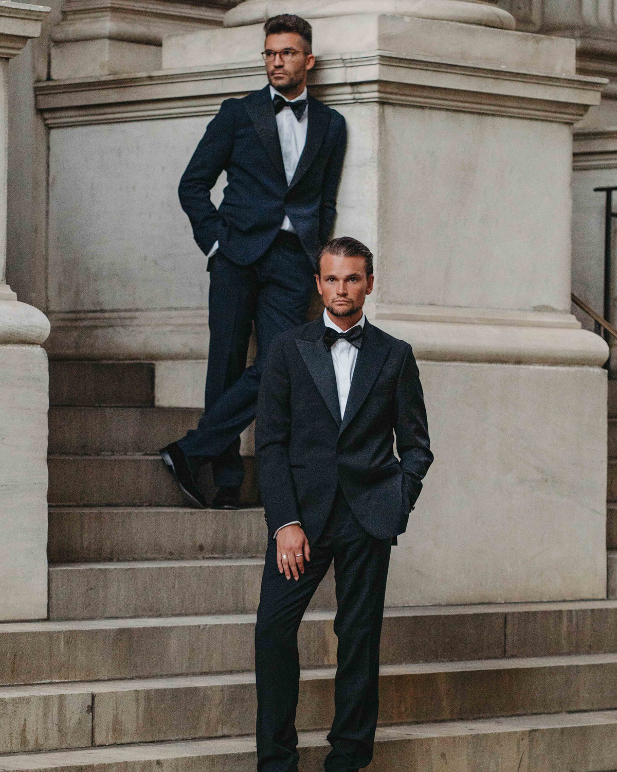 Two men in black tuxedos with white shirts and black bow ties pose on the stone steps of a grand classical building.