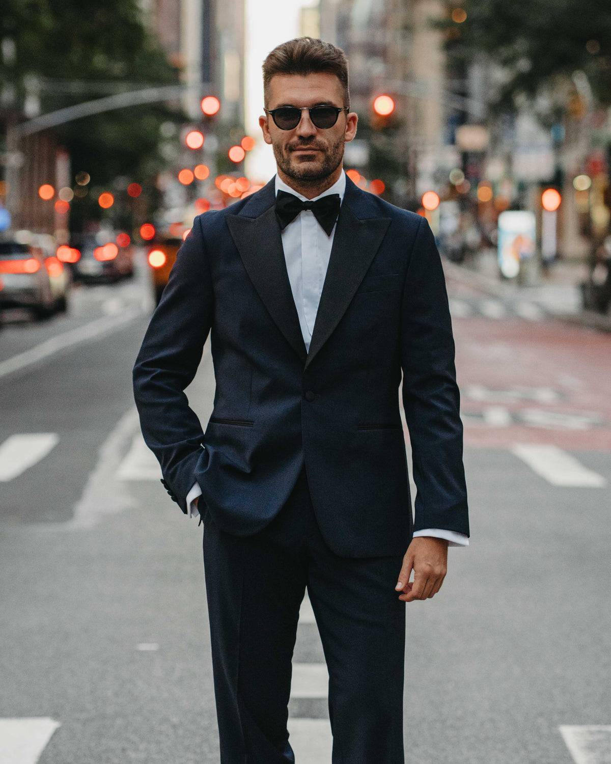 A man wearing a dark navy blue tuxedo with black satin lapels, a white dress shirt, and a black bow tie stands on a city street at dusk.