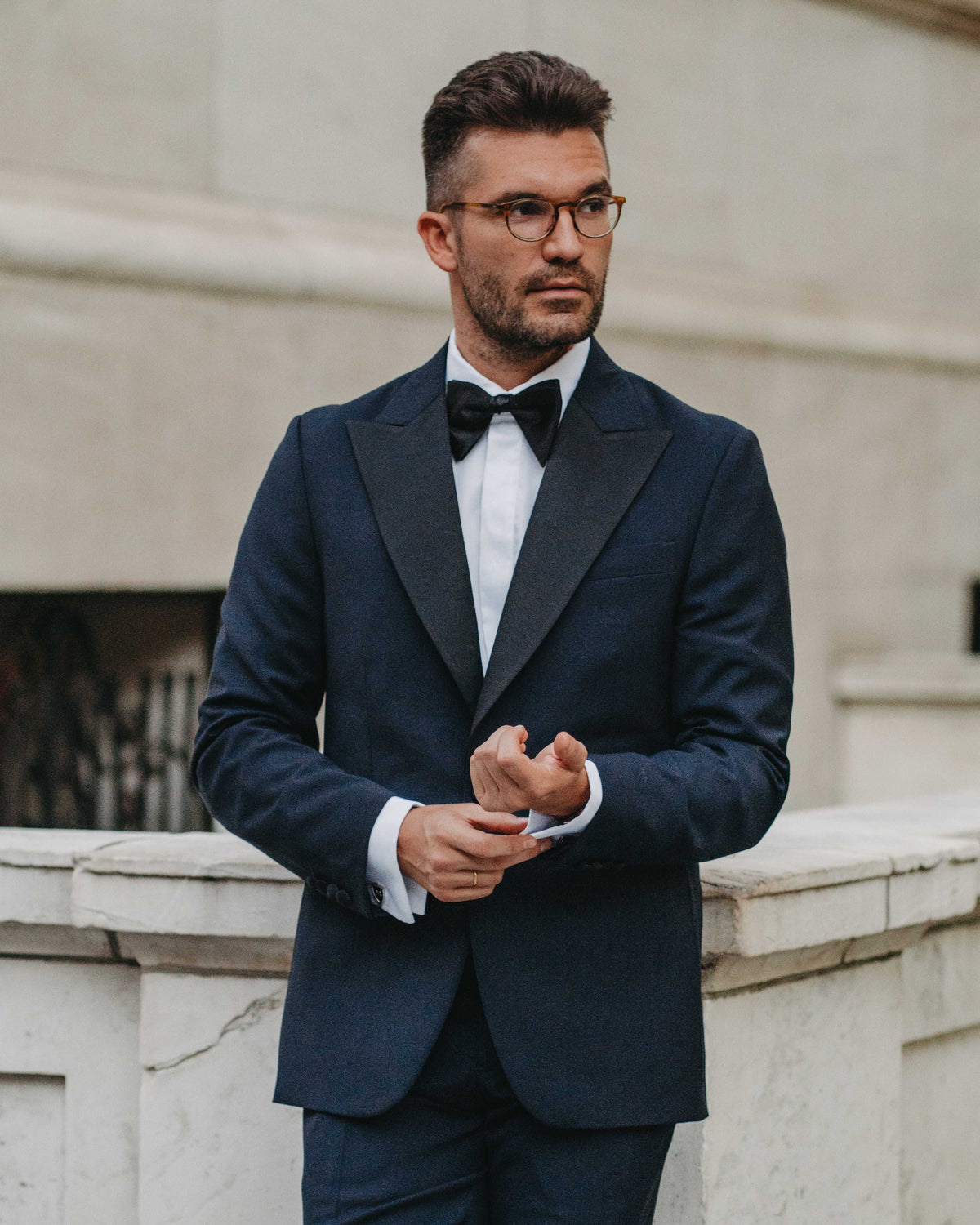 A man wearing a dark navy blue tuxedo jacket with black satin lapels, a white dress shirt, and a black bow tie, standing outdoors against a light stone building.