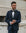 A man wearing a dark navy blue tuxedo jacket with black satin lapels, a white dress shirt, and a black bow tie, standing outdoors against a light stone building.