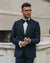 A man wearing a dark navy blue tuxedo jacket with black satin lapels, a white dress shirt, and a black bow tie, standing outdoors against a light stone building.