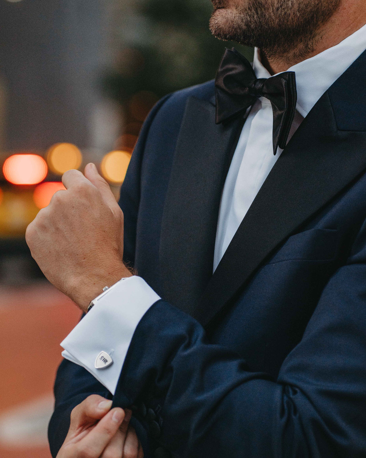 A close-up lifestyle shot of a man in a navy blue tuxedo jacket with black satin lapels, a white dress shirt, a black bow tie, and white cufflinks, adjusting his cufflink in an outdoor setting with blurred city lights.