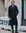 A man wearing a long black double-breasted overcoat, blue dress shirt, blue patterned tie, and dark trousers stands on a city sidewalk in front of a stone building.