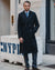 A man wearing a long black double-breasted overcoat, blue dress shirt, blue patterned tie, and dark trousers stands on a city sidewalk in front of a stone building.