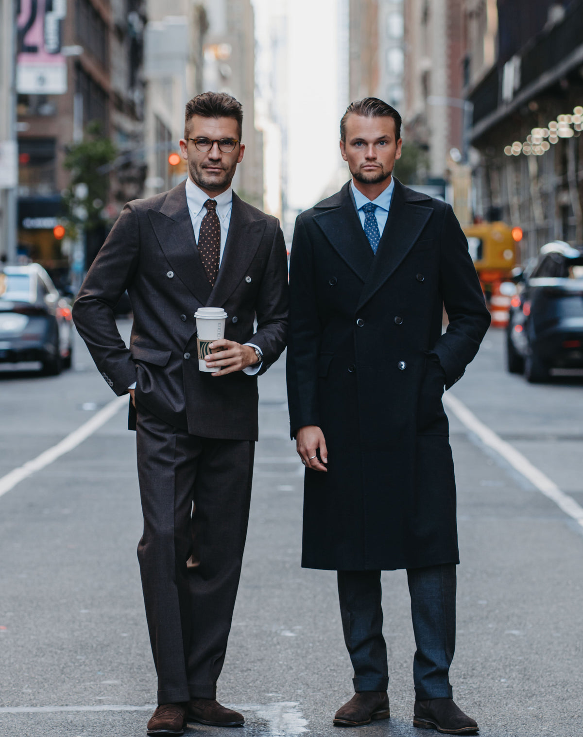 Two men standing on a city street wearing dark tailored outerwear and suits, with one holding a takeaway coffee cup. The scene includes buildings, cars, and road markings in a real urban environment.