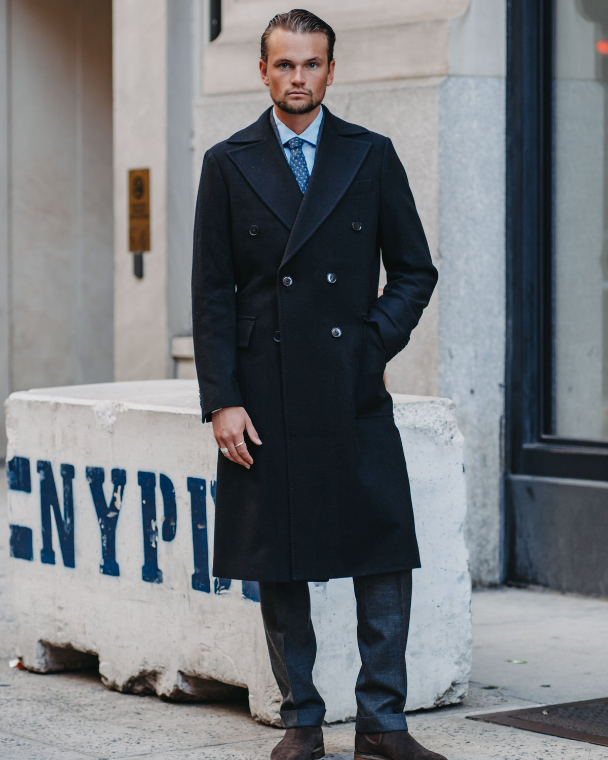 A man wearing a long black double-breasted overcoat, blue dress shirt, blue patterned tie, and dark trousers stands on a city sidewalk in front of a stone building.