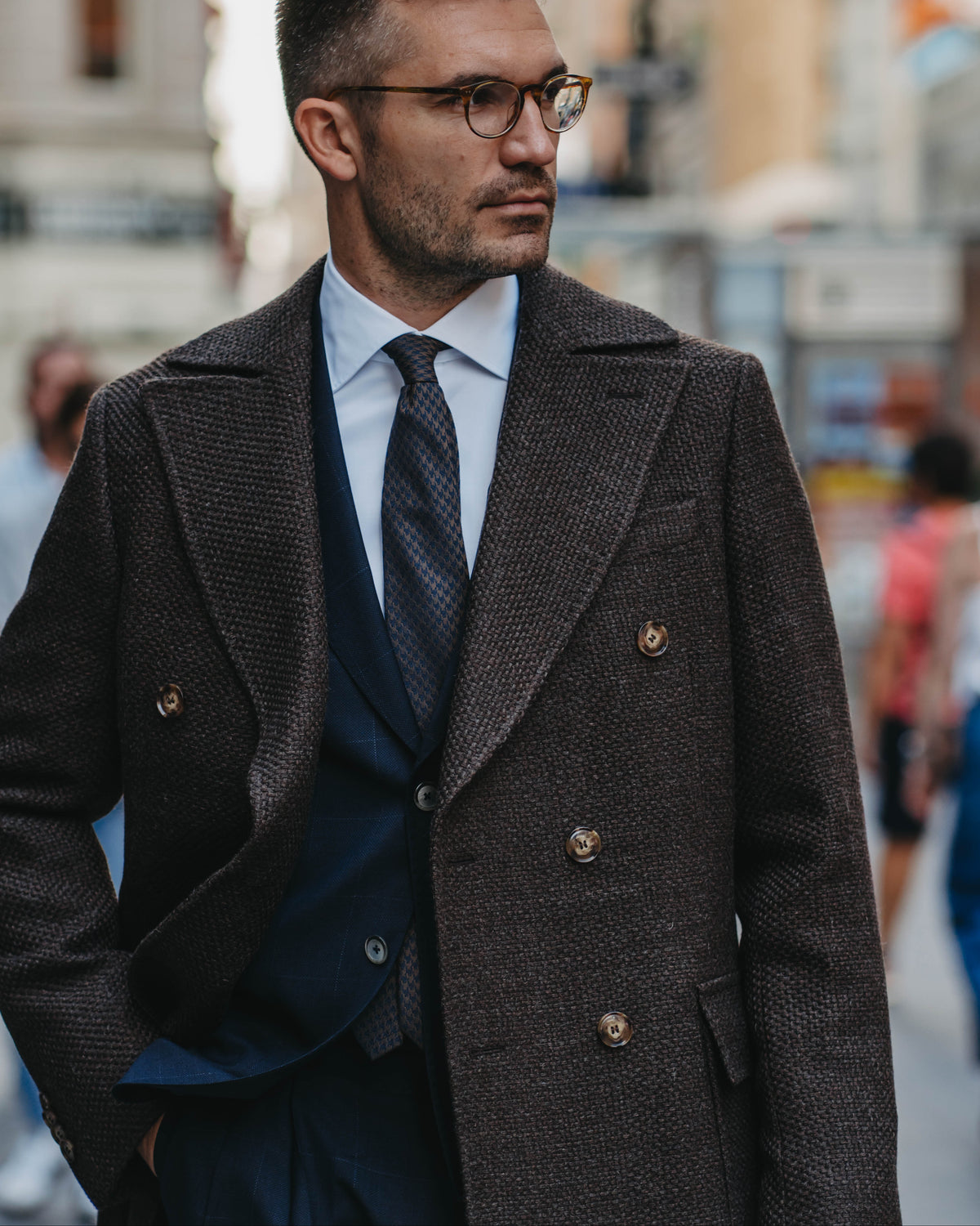 A man wearing a dark brown textured double-breasted overcoat, navy blue suit, white shirt, and patterned tie, standing on a blurred city street.
