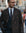 A man wearing a dark brown textured double-breasted overcoat, navy blue suit, white shirt, and patterned tie, standing on a blurred city street.