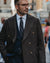 A man wearing a dark brown textured double-breasted overcoat, navy blue suit, white shirt, and patterned tie, standing on a blurred city street.