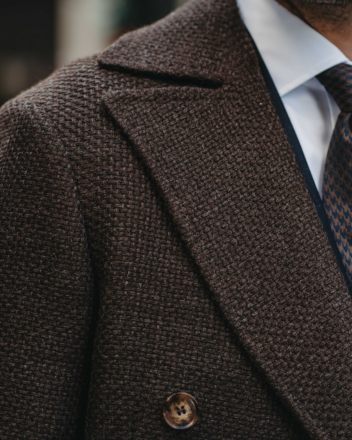 Close-up of a man wearing a dark brown textured double-breasted overcoat, a white dress shirt, and a patterned tie.