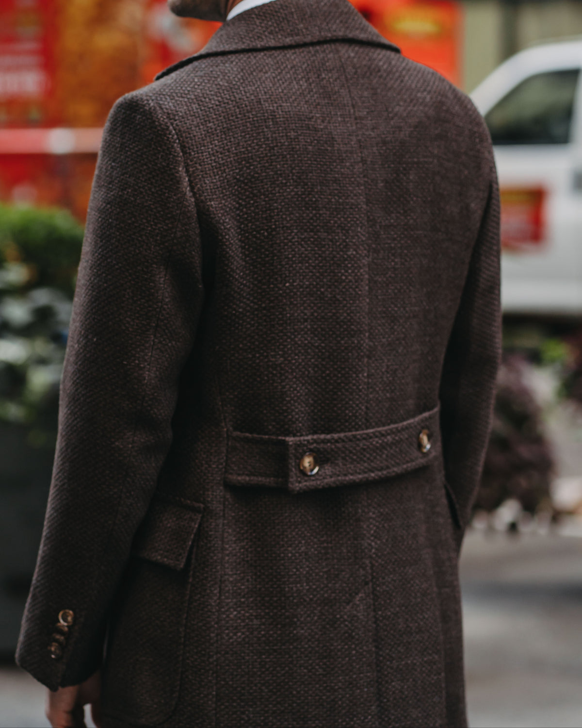 A man viewed from behind wearing a dark brown textured wool overcoat with a matching belt and gold-tone buttons, set against a blurred city street background.