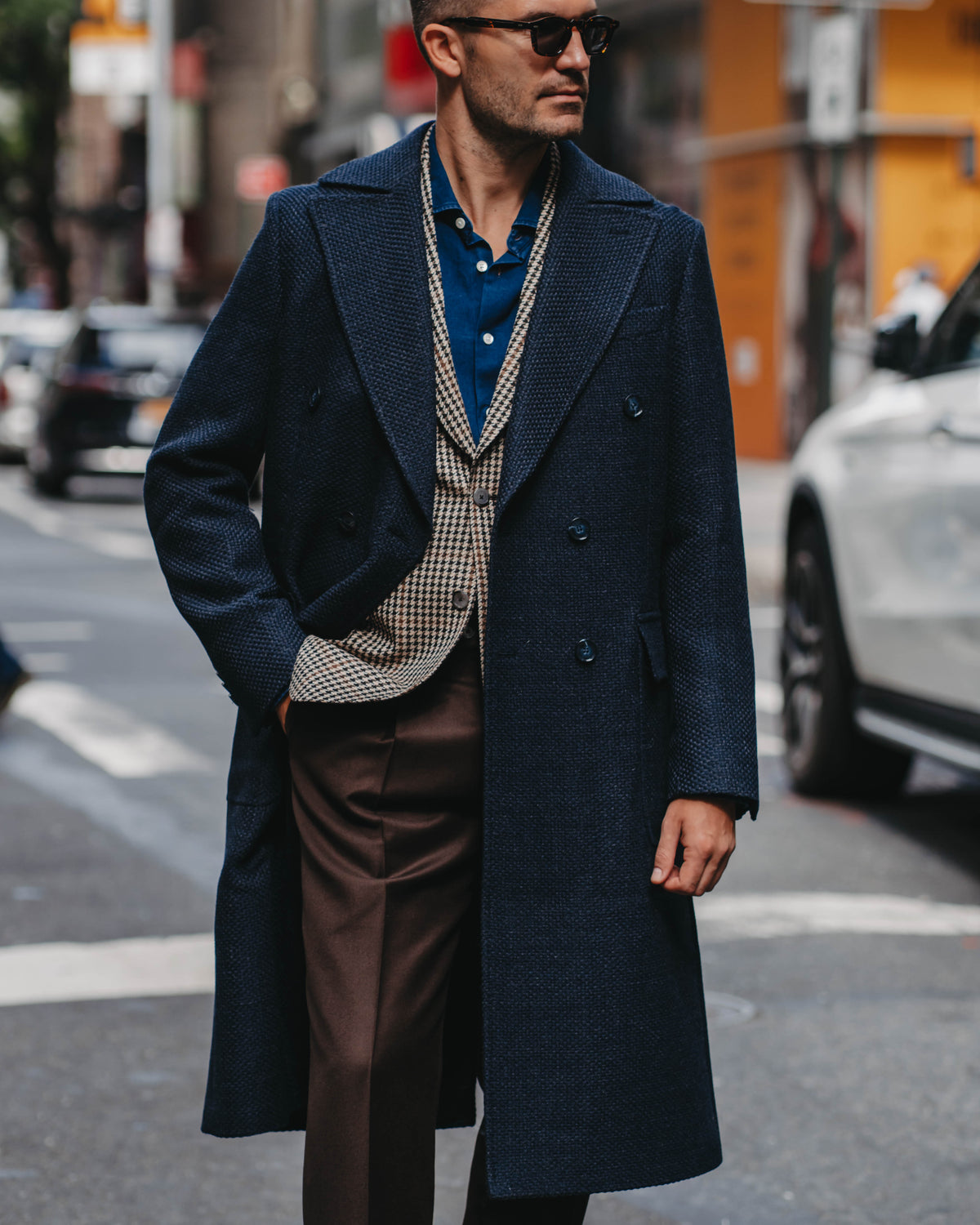A man wearing a long navy blue overcoat, a blue polo shirt, a beige and black gingham sweater vest, brown trousers, and sunglasses, walking on a city street.