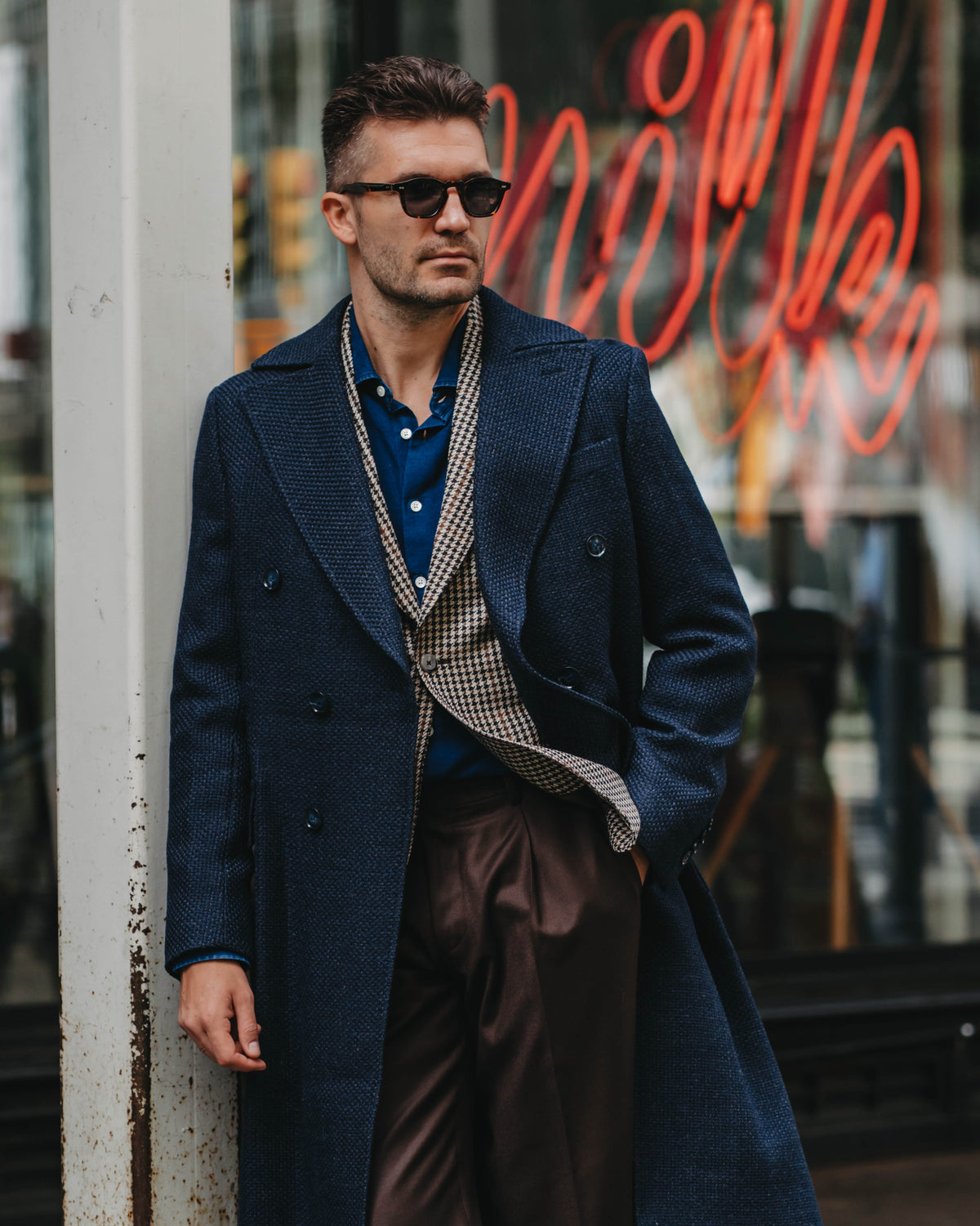 A man wearing a dark navy double-breasted overcoat, a patterned vest over a blue shirt, and brown trousers, leaning against a weathered white column in front of a storefront with a red neon sign.
