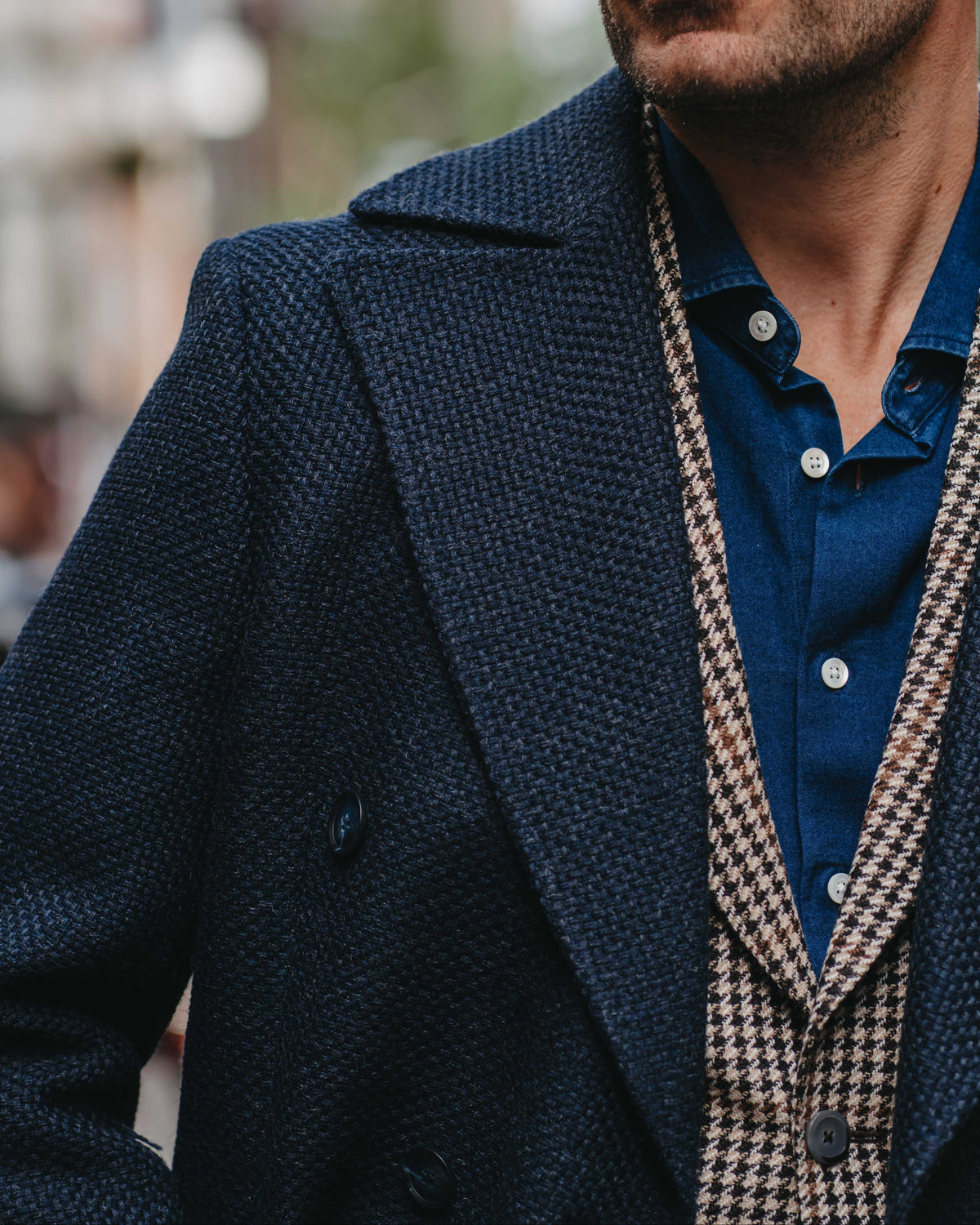 A close-up of a man wearing a dark navy textured double-breasted coat over a blue button-down shirt and a beige-and-black houndstooth suit jacket, with dark sunglasses.
