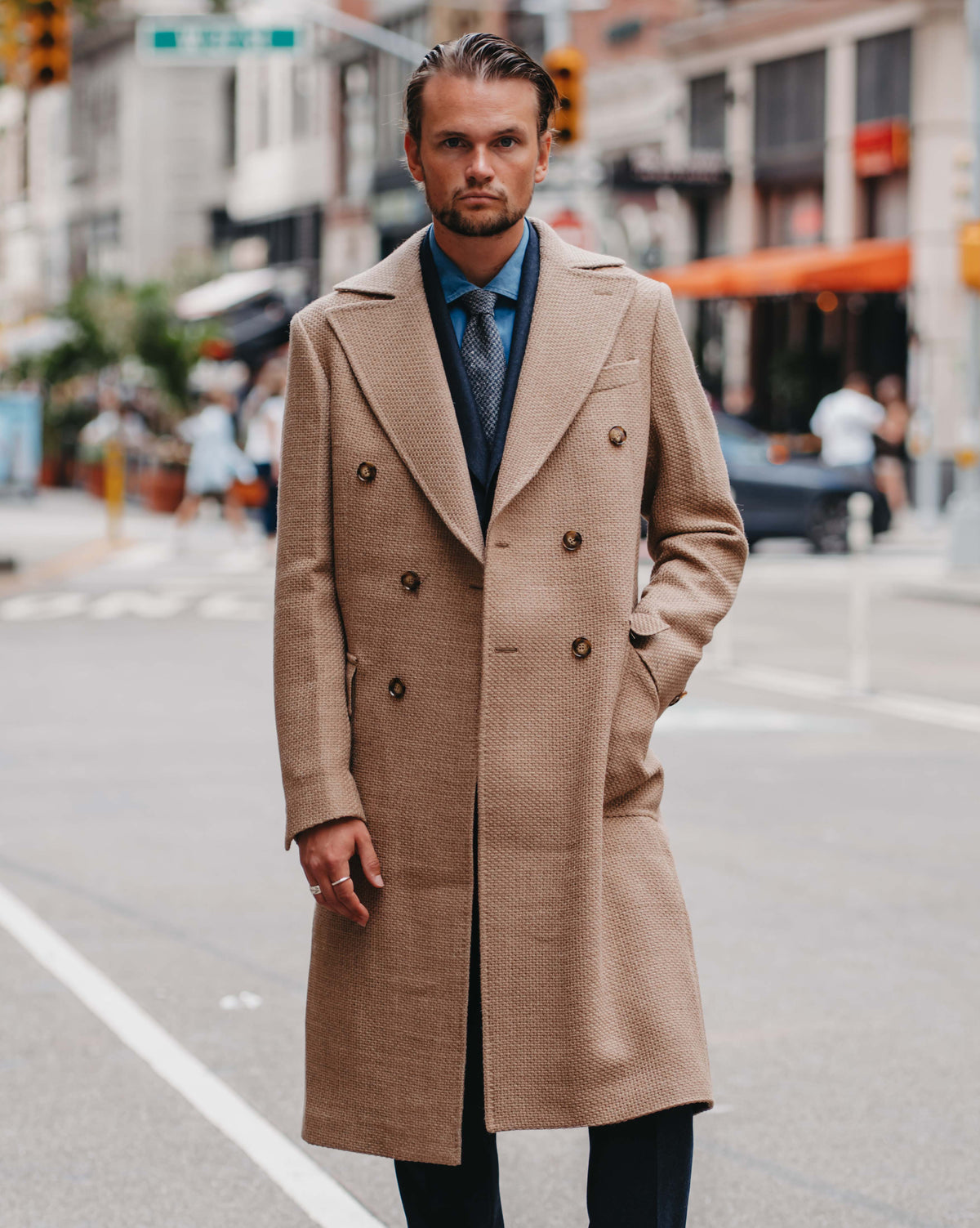A man stands on a city street wearing a long beige double-breasted overcoat over a blue shirt and dark trousers, with brown shoes. The background shows a blurred urban street scene with buildings, pedestrians, and traffic lights.