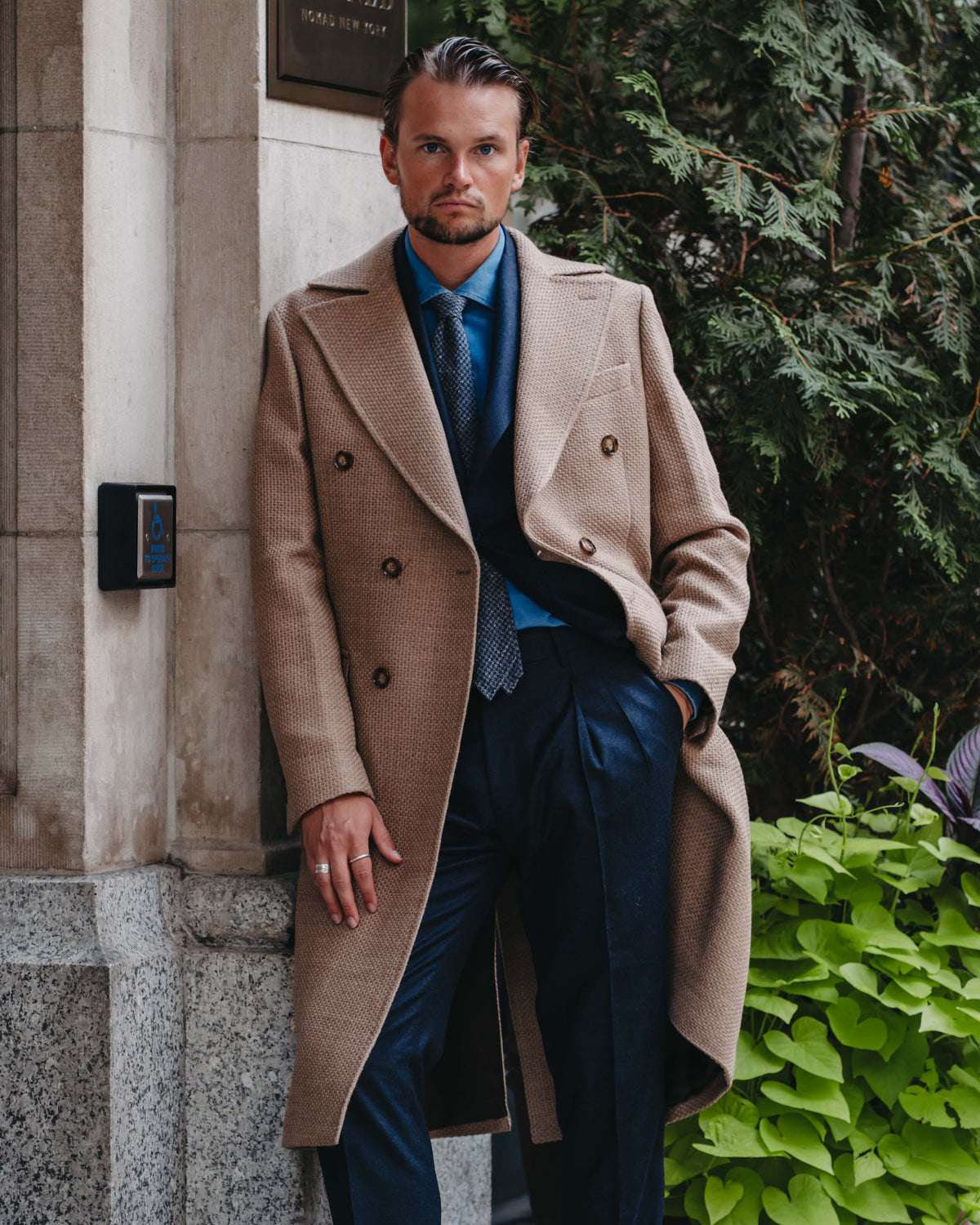 A man wearing a beige double-breasted overcoat, navy trousers, a blue shirt, and a blue tie leans against a stone building exterior with green foliage in the background.