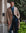 A man wearing a beige double-breasted overcoat, navy trousers, a blue shirt, and a blue tie leans against a stone building exterior with green foliage in the background.
